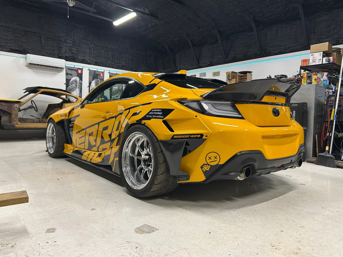 Error 404 Motorsports Toyota GR86 widebody build — yellow livery with carbon fiber trunk spoiler, rear three-quarter view